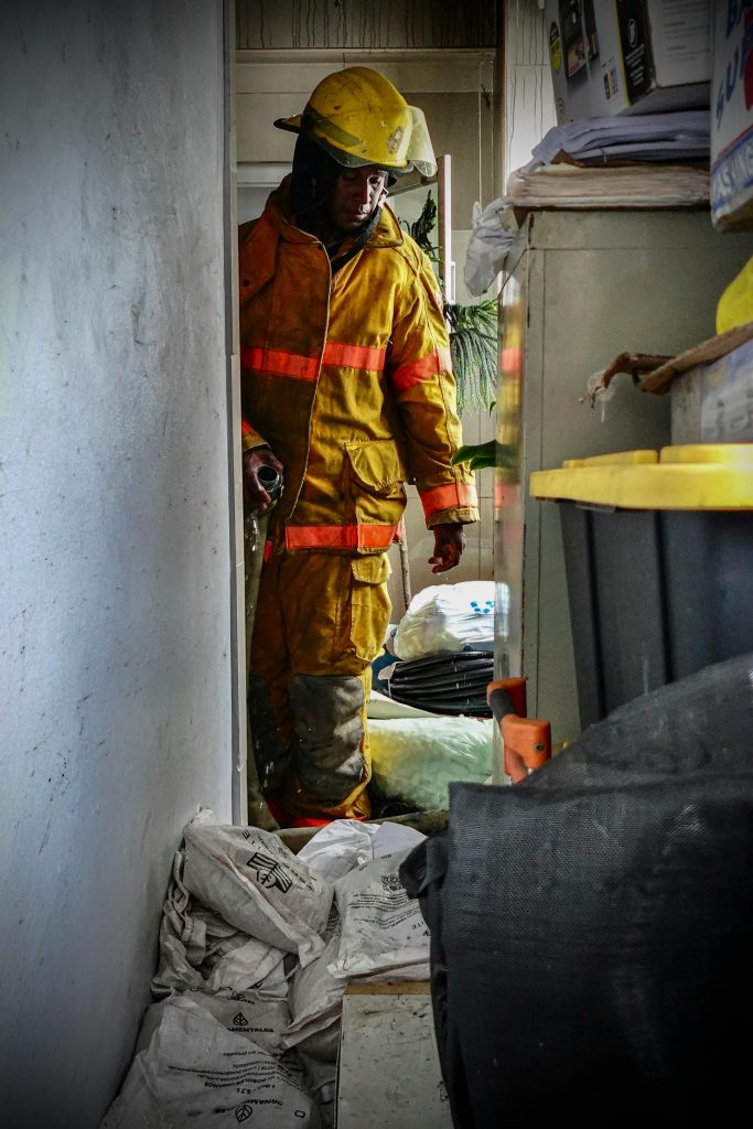 A firefighter in a yellow uniform inspects a cluttered room. Indoor scene, safety equipment visible.